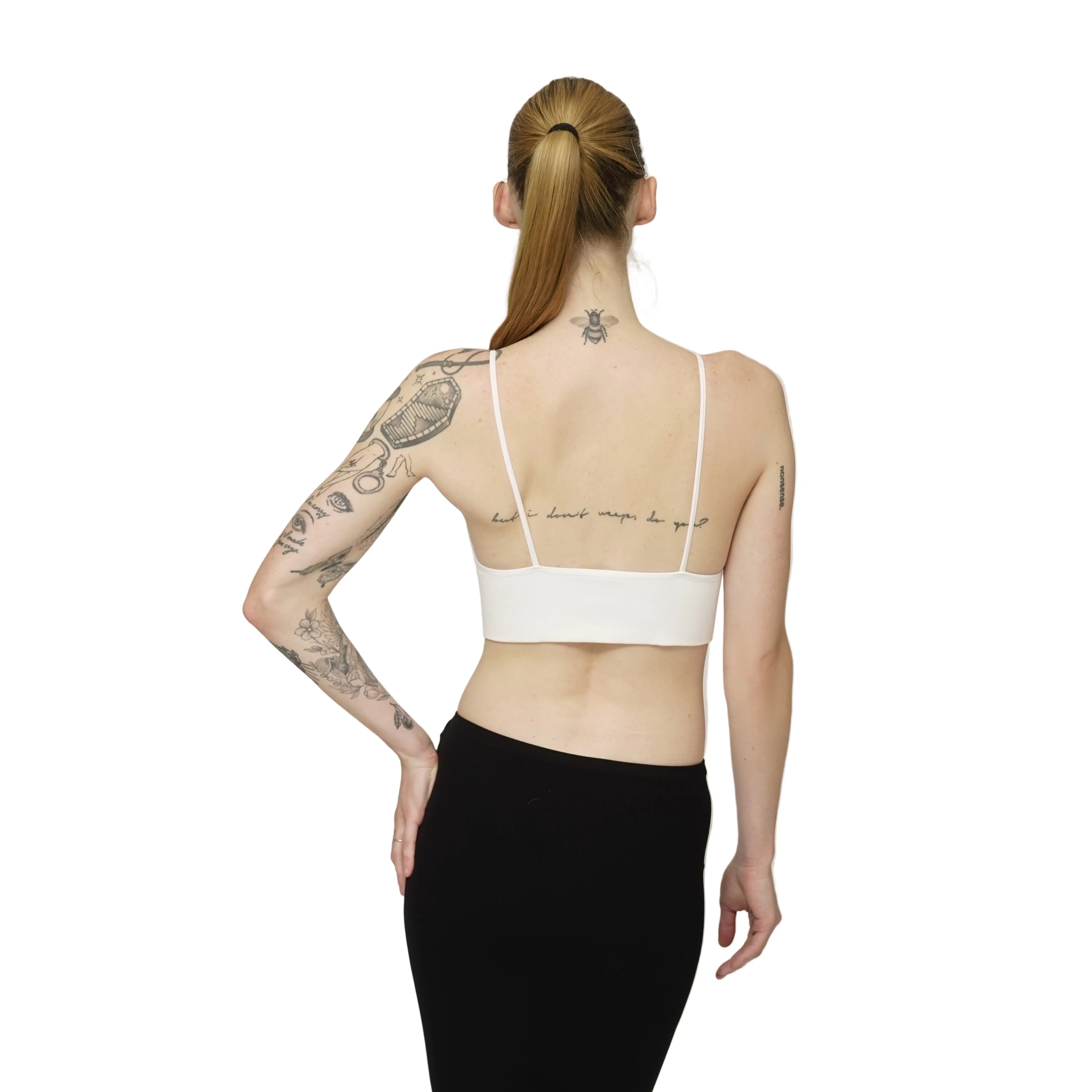 Person wearing a white bralette with tattoos on arms and back, standing against a plain background
